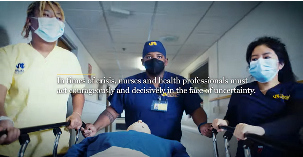 Two women, one Black, and one Black man wearing Drexel's blue and yellow scrubs pushing a hospital bed with a simulation manikin down a long corridor.